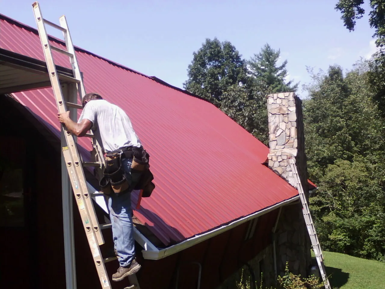 Roofer on a ladder beside a red metal roof and stone chimney — Soffit & Fascia Repair by Roofing Brothers in Lesterville